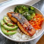 Close-up of a delicious salmon rice bowl featuring a glazed salmon fillet, sliced avocado, steamed broccoli, and shredded carrots over white rice.