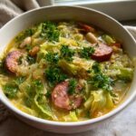 Close-up of a bowl of hearty cabbage soup featuring sliced smoked sausage, white beans, and fresh parsley garnish.
