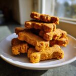 A stack of freshly baked, orange-colored, bone-shaped dog cookies resting on a white plate.