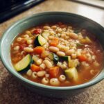 Close-up of a steaming bowl of minestrone soup filled with ditalini pasta, white beans, zucchini, and carrots.