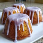 Four golden mini lemon cakes with a thick, pale purple lavender glaze dripping down the sides, served on a white plate.