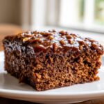 A close-up of a rich, dark slice of Dr Pepper cake topped with a glossy caramel-pecan glaze.