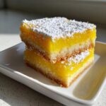 Two stacked squares of bright yellow no-bake lemon bars, dusted generously with white powdered sugar, resting on a white dish.
