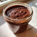Close-up of rich, thick chocolate overnight oats mixture in a clear glass jar, illuminated by sunlight.