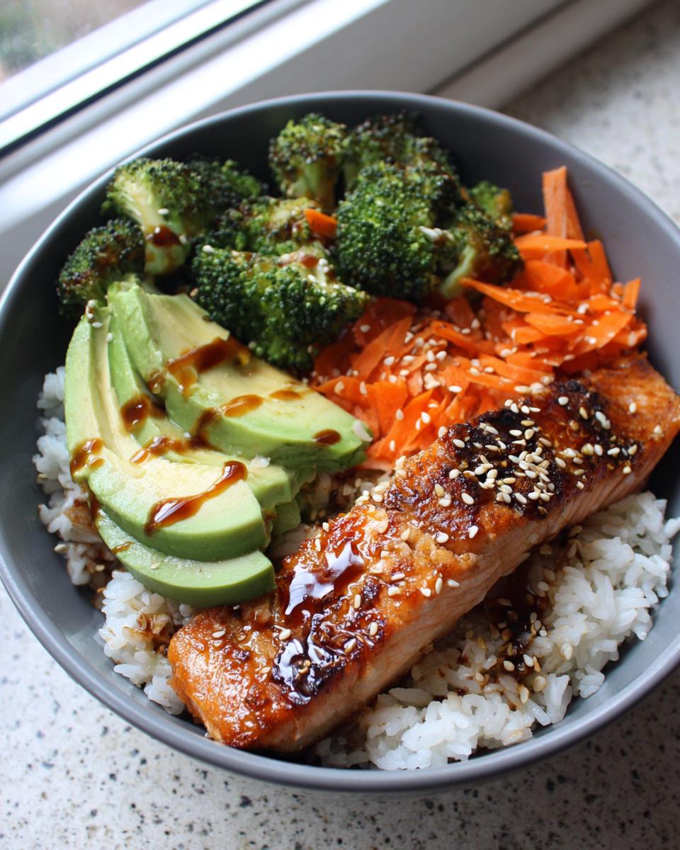 Close-up of a perfectly cooked salmon rice bowl topped with glazed salmon, avocado, roasted broccoli, and shredded carrots.