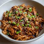 Close-up of shredded crockpot teriyaki chicken coated in thick sauce, topped with sesame seeds and green onions.