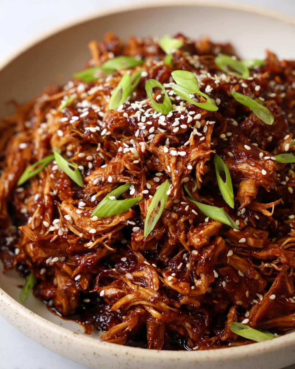 Close-up of shredded crockpot teriyaki chicken coated in dark sauce, topped with sesame seeds and green onions.