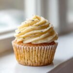 A single, beautifully frosted lavender honey cupcake topped with dried lavender buds sitting on a white surface.