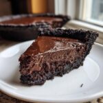 A decadent slice of chocolate fudge pie with a thick, dark, crumbly crust resting on a white plate.