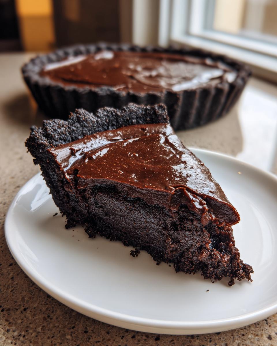 A thick slice of dark chocolate fudge pie with a glossy top sits on a white plate, with the rest of the chocolate fudge pie blurred in the background.