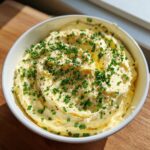 A bowl of fluffy, yellow smoked cream cheese dip garnished generously with freshly chopped chives and black pepper.