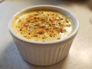 A small white ramekin filled with creamy, yellow spicy caesar dressing, topped with blue cheese crumbles and paprika.