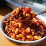 A spoonful of rich american chop suey, featuring elbow macaroni coated in a hearty meat and tomato sauce, lifted from a white bowl.
