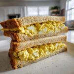Two halves of a thick egg salad sandwich stacked on top of each other, showing creamy yellow filling.