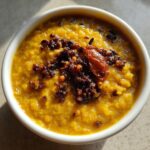 A close-up overhead shot of a white bowl filled with bright yellow dhal, topped with a dark, spicy tempering (tadka).