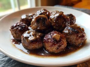 A close-up of several browned venison meatballs smothered in rich Marsala sauce with sliced mushrooms, served on a white plate.
