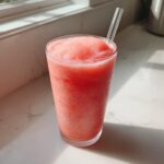 A tall glass filled with a bright pink, icy watermelon cloud slushie, featuring a clear straw, sitting on a sunny white countertop.