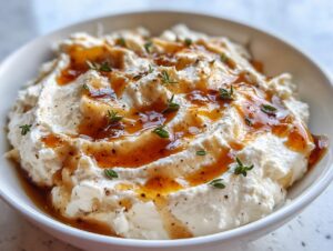 Close-up of a creamy, white ricotta dip topped with golden honey drizzle, black pepper, and fresh thyme sprigs.