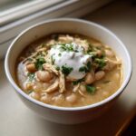 A close-up of a white bowl filled with creamy chicken chili recipe, topped with shredded chicken, white beans, sour cream, and fresh parsley.