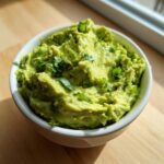 Close-up of a white bowl filled with chunky, bright green Authentic Guacamole, garnished with chopped green onions and cilantro.