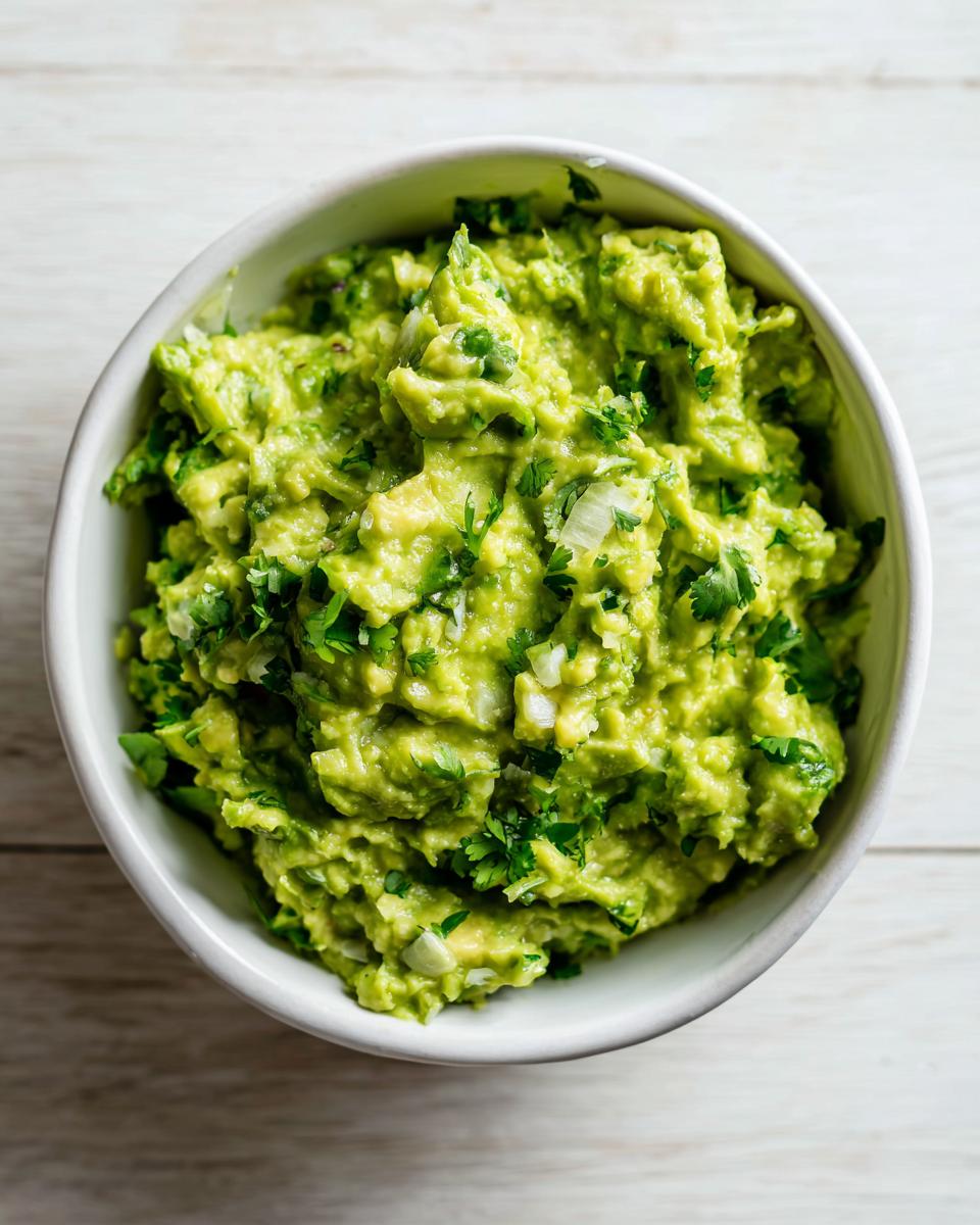 Overhead view of a white bowl filled with chunky, bright green Authentic Guacamole topped with cilantro and onion.