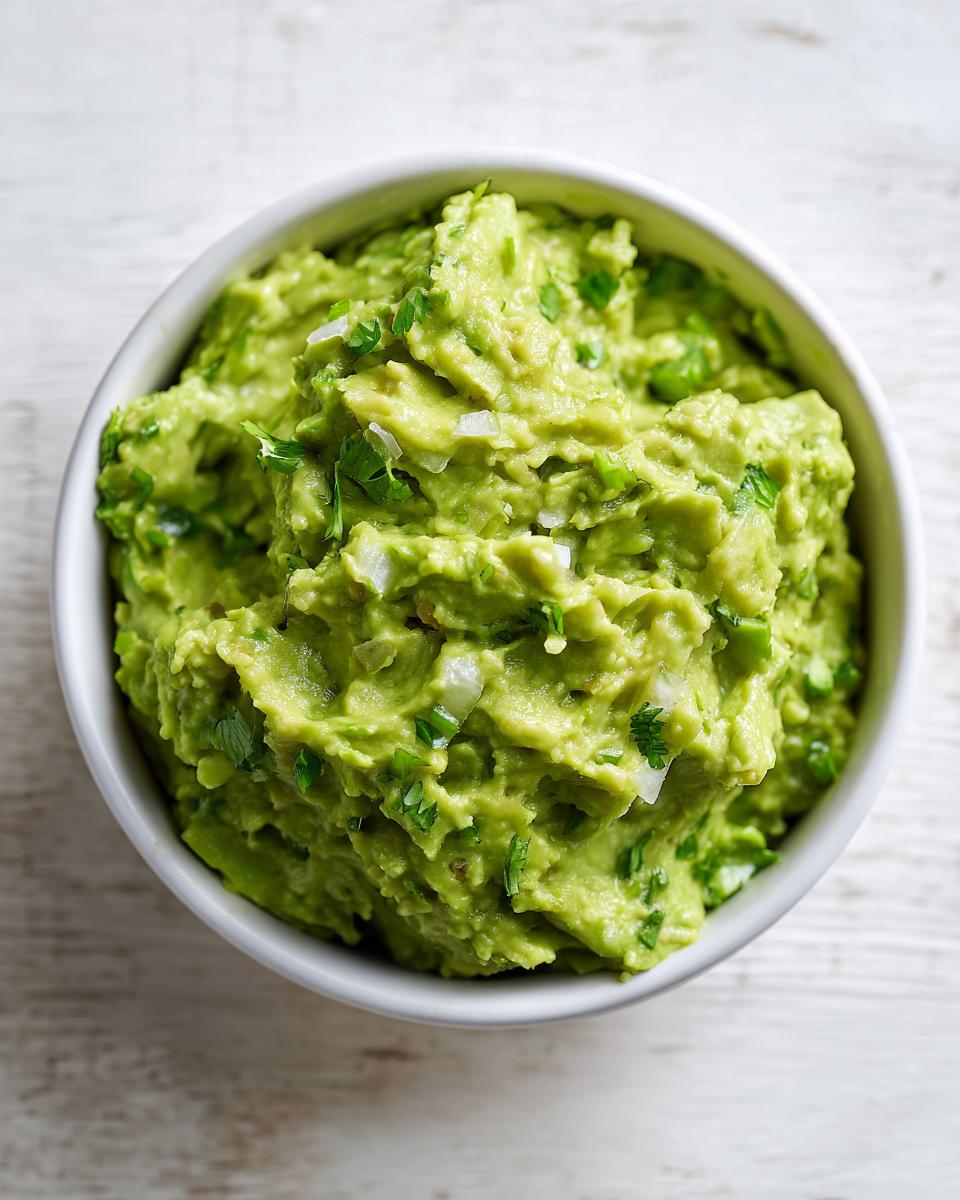 Overhead view of a white bowl filled with chunky, bright green authentic guacamole, visible pieces of onion and cilantro.