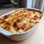 A close-up of a freshly baked Artichoke cream dip in a white casserole dish, featuring a bubbly, golden-brown melted cheese crust.