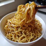 Close-up of creamy Carbonara buldak ramen noodles being lifted from a white bowl with dark chopsticks, showing chili flakes.