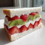 A cross-section view of a vibrant Japanese fruit sandwich filled with layers of fresh strawberries, kiwi slices, and whipped cream.