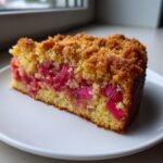 A close-up of a moist slice of Rhubarb Coffee Cake featuring bright pink rhubarb pieces and a golden brown crumble topping.