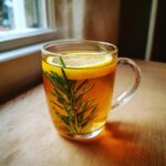 A clear glass mug filled with warm rosemary tea, featuring a fresh rosemary sprig and a lemon slice floating on top.