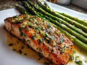 Close-up of a juicy fillet of sheet pan garlic butter salmon topped with herbs and resting in butter sauce next to roasted asparagus.