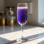 A tall flute glass filled with a vibrant, sparkling purple Blueberry Lemosas cocktail sitting on a sunlit marble counter.
