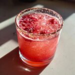Close-up of a vibrant, icy Strawberry mezcal margarita in a rocks glass with a colorful salt rim.
